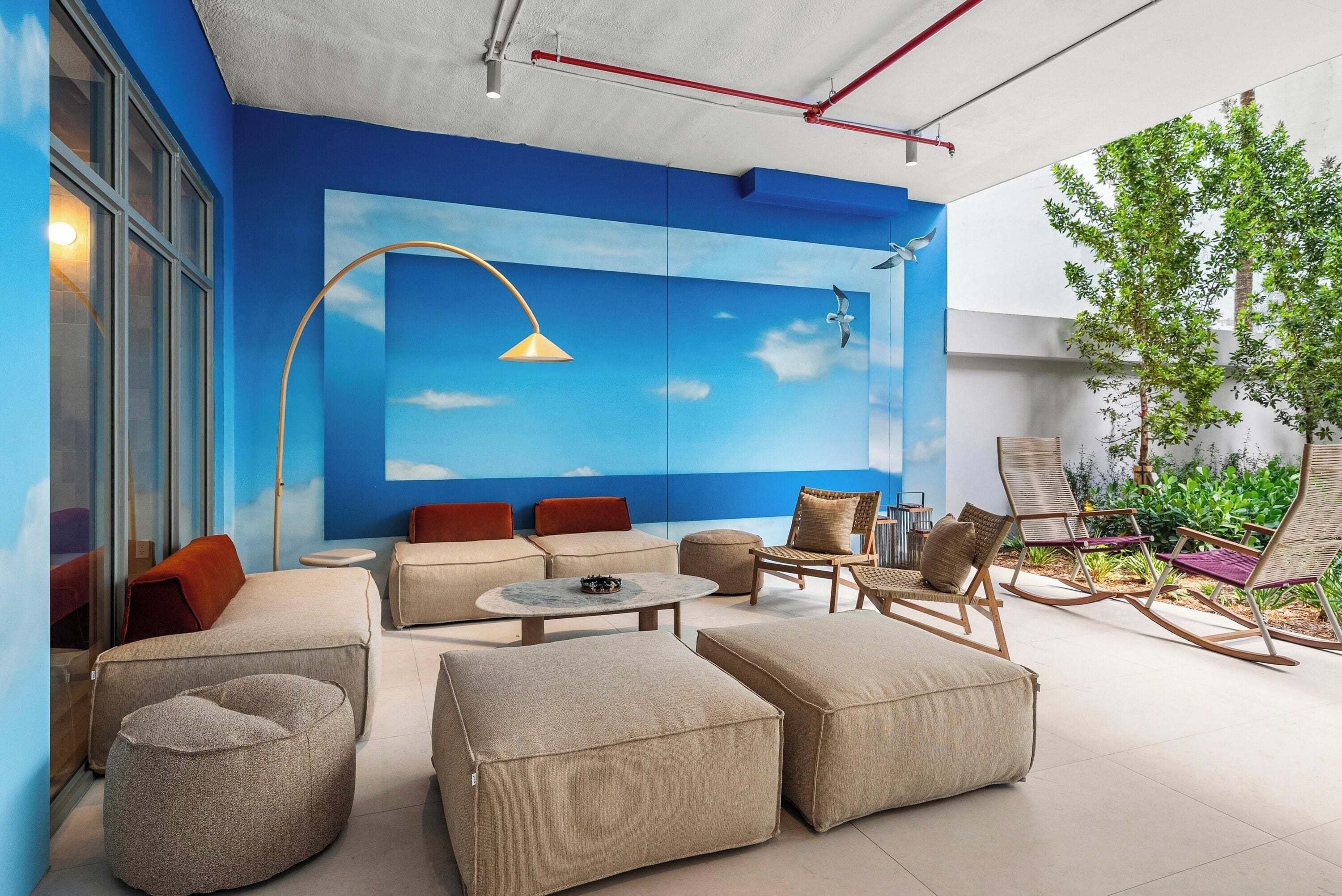 A modern outdoor seating area with sofas, ottomans, and rocking chairs, a round table, and a mural of a blue sky with clouds.