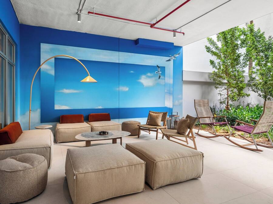 A modern outdoor seating area with sofas, ottomans, and rocking chairs, a round table, and a mural of a blue sky with clouds.
