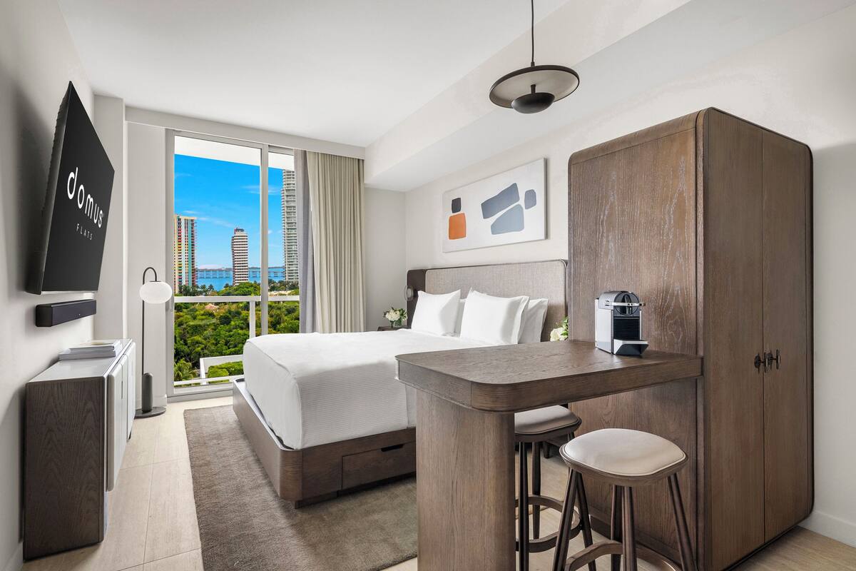 Habitación de hotel moderna con cama, escritorio, silla, armario, televisor y vista a la ciudad.