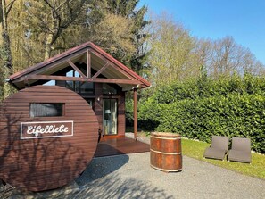A wooden cabin with a sign reading "Eifelliebe," a wooden barrel, and two lounge chairs.