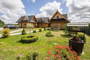 Une cabane en rondins avec un toit en pente, une véranda en bois et un jardin bien entretenu, agrémenté d’arbustes et de fleurs.