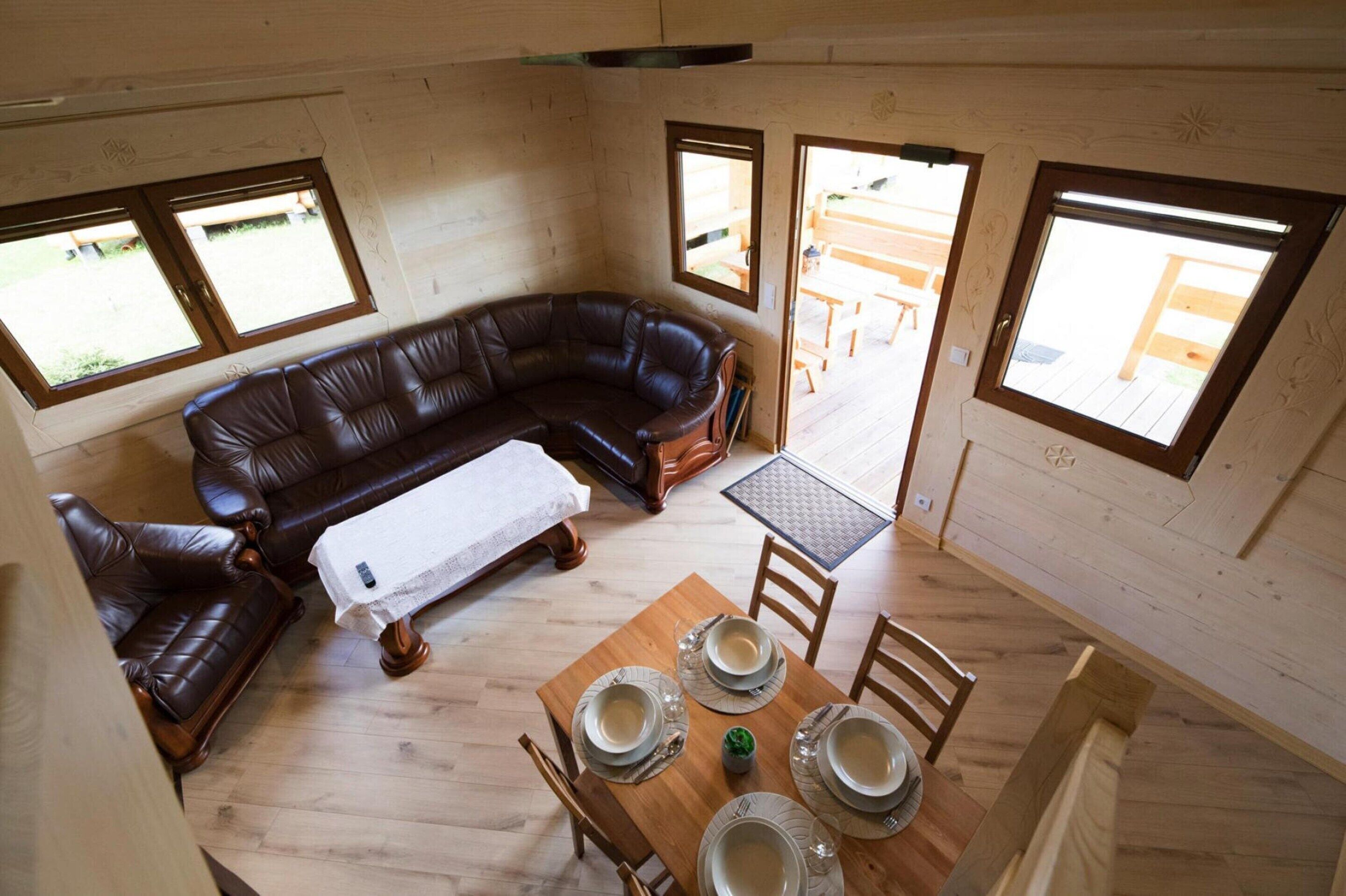 A wooden interior with a brown leather sofa, a dining table set with plates, and two windows providing natural light.