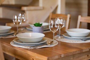 Une table de salle à manger en bois, dressée avec de l’assiette blanche, des bols et des verres à vin, agrémentée d’une petite plante en pot.