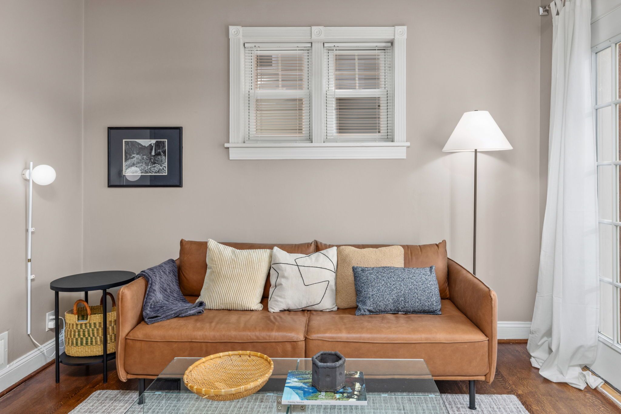 A living room with a brown leather sofa, a glass coffee table, a framed picture on the wall, a floor lamp, and a window with white blinds.