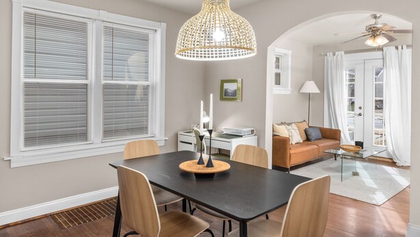 Uma sala de jantar com mesa preta, cadeiras de madeira, luminária pendente trançada e ventilador de teto branco.