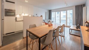A modern kitchen with white cabinets, a built-in oven, and a dining area with a wooden table and chairs. - Knokke City Centre Apartment for 6 (Knokke-Heist)
