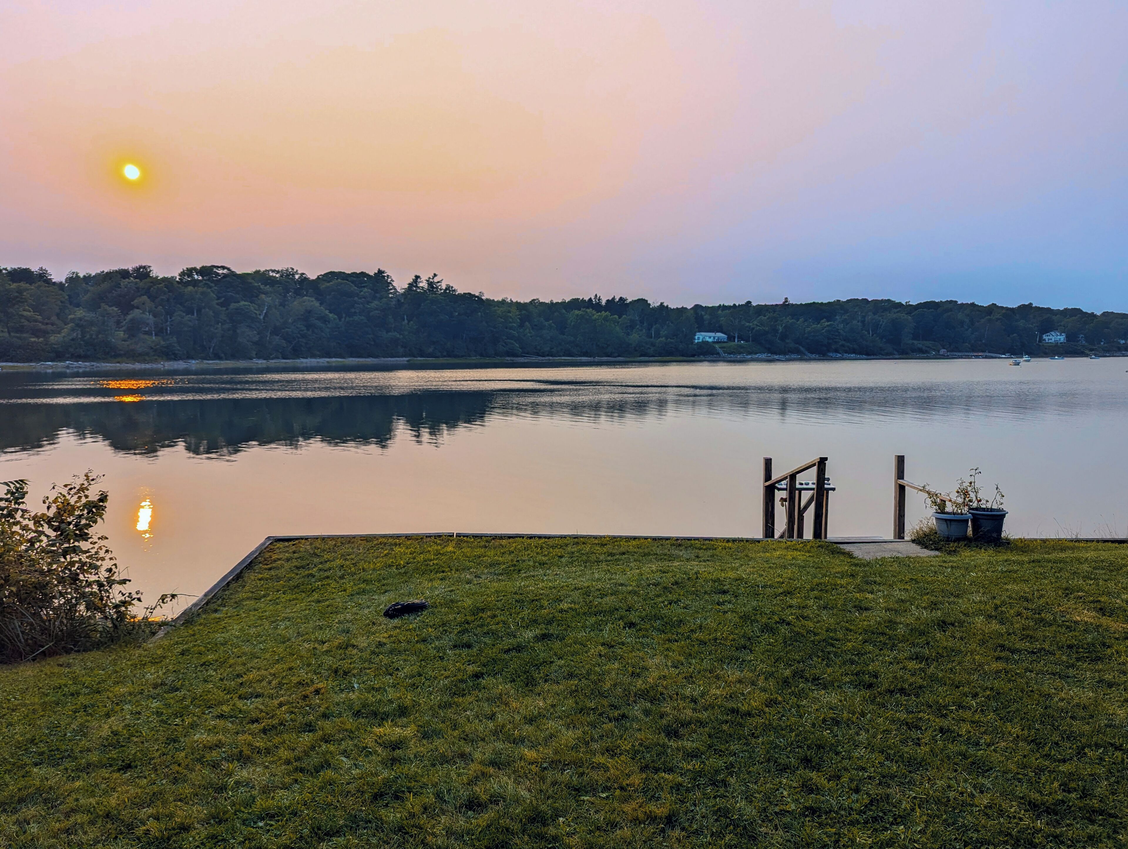 A calm lake at sunset with a grassy area in the foreground and a dock extending into the water.