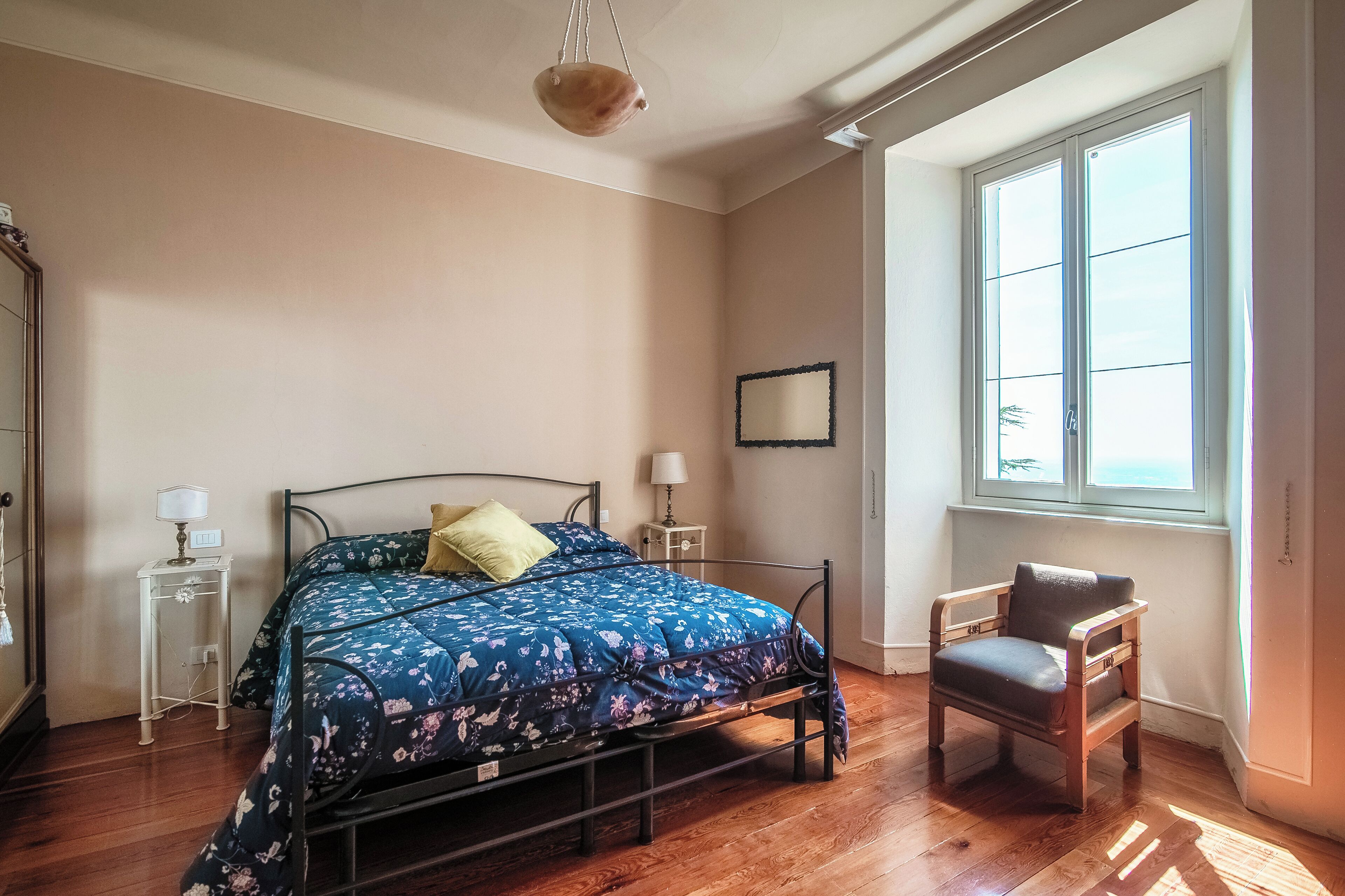 Un dormitorio con cama, mesitas de noche, una silla, un ventanal y una lámpara colgante.