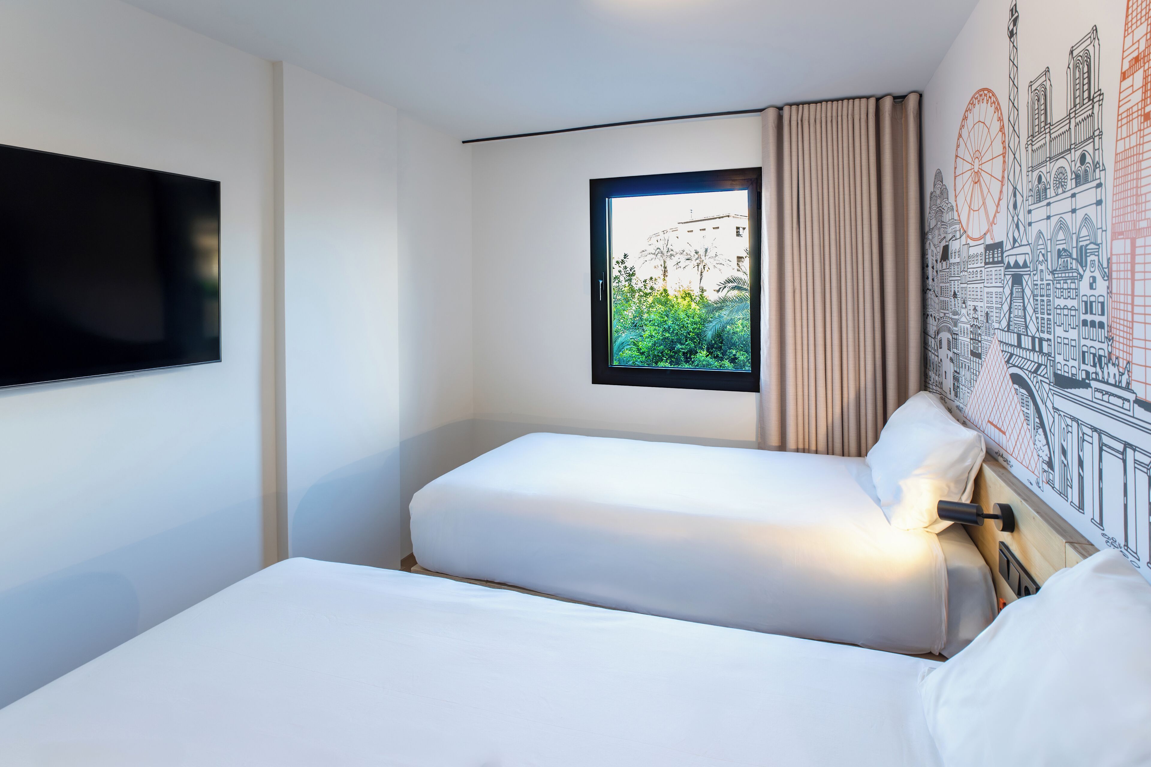 A hotel room with two beds, a TV, a window with curtains, and a cityscape mural on the wall.