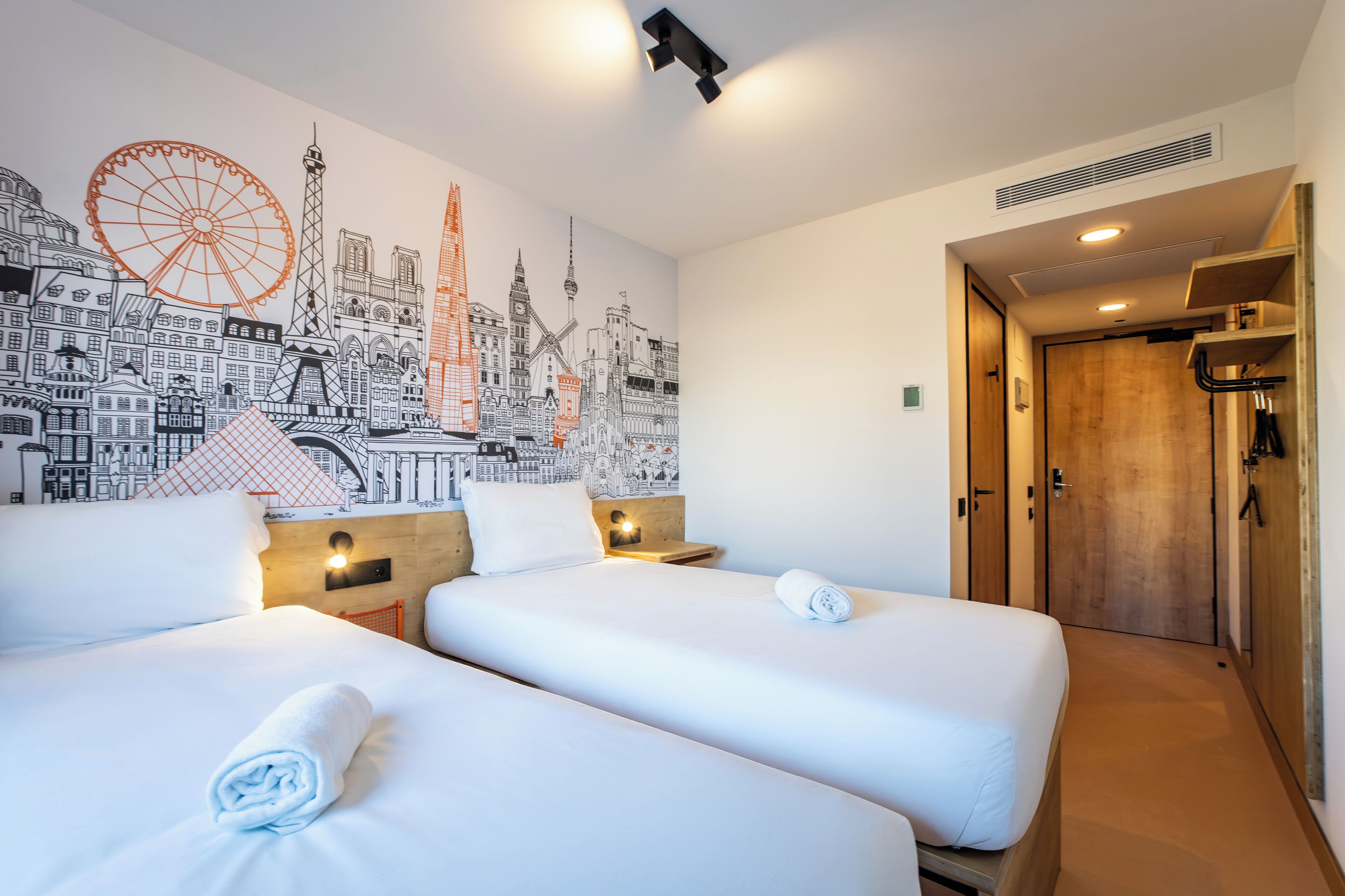 A hotel room with two beds, a mural of cityscapes, a wooden door, and a small table with a lamp.