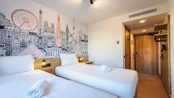 A hotel room with two beds, a mural of cityscapes, a wooden door, and a small table with a lamp.