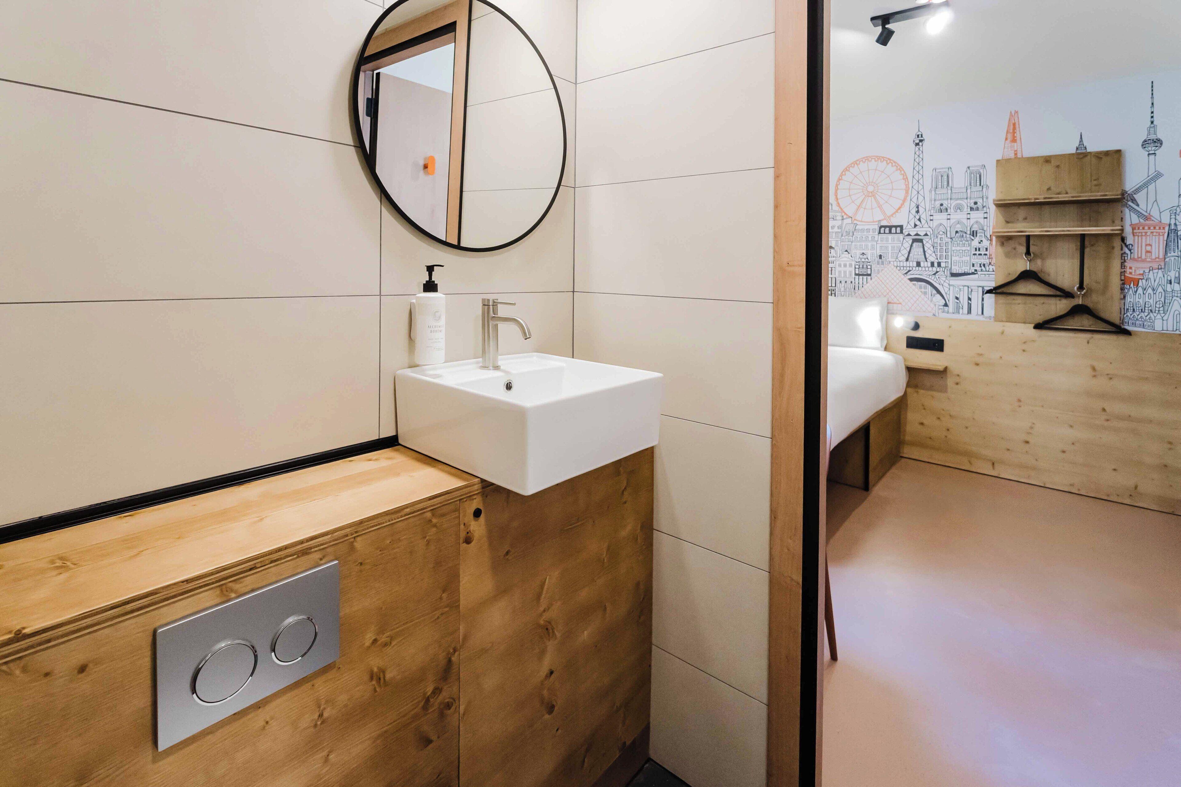 Un baño moderno con un tocador de madera, un espejo redondo, un lavamanos blanco y una pared con un mural de un paisaje urbano.