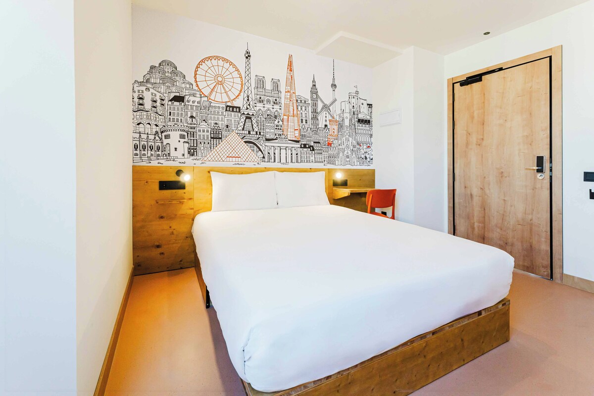 A hotel room with a large bed, a wooden headboard, a cityscape mural on the wall, a wooden door, and a nightstand with a lamp.