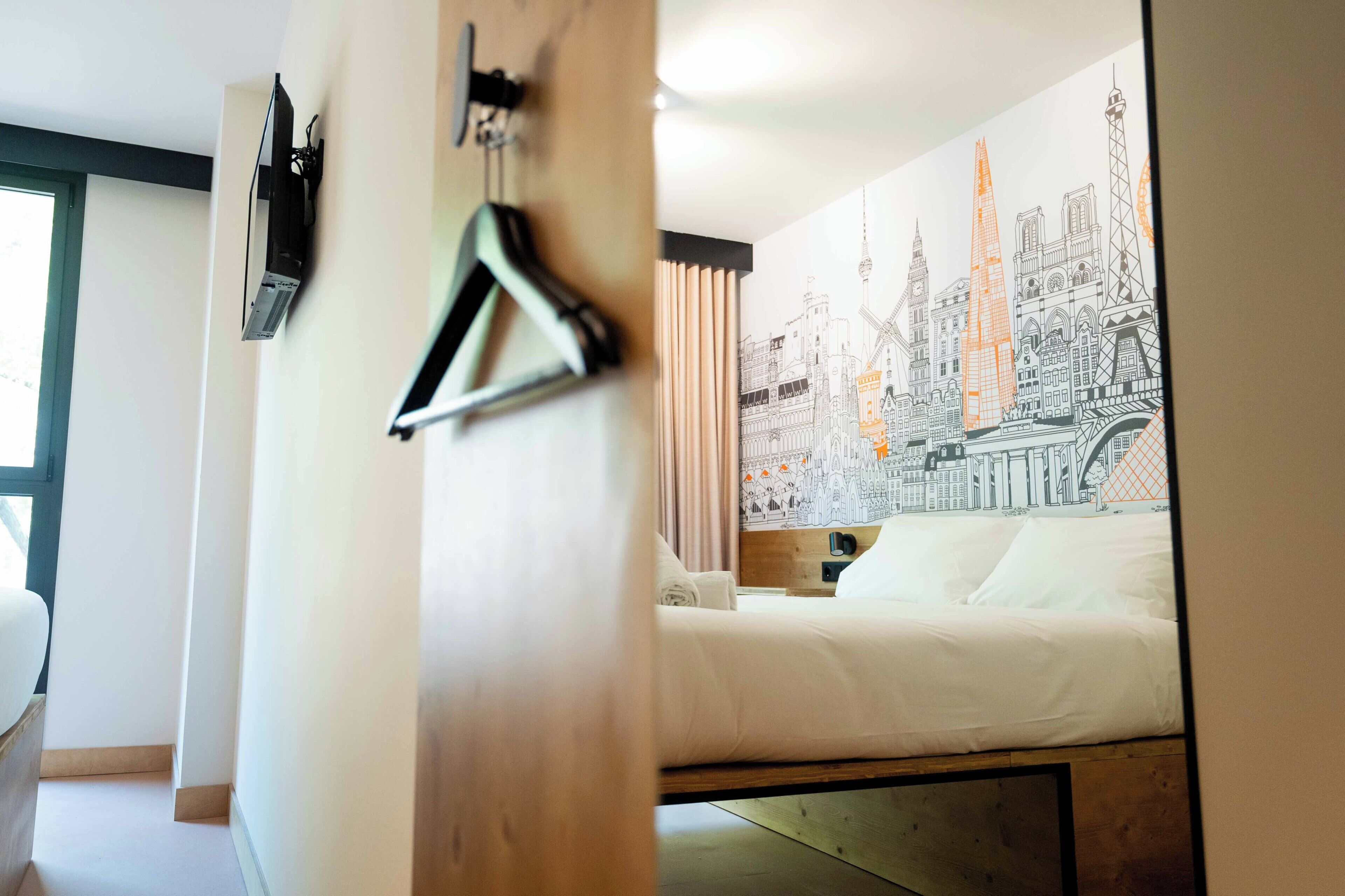 A hotel room with a bed, a wall with a cityscape mural, a hanging coat rack, and a window with curtains.