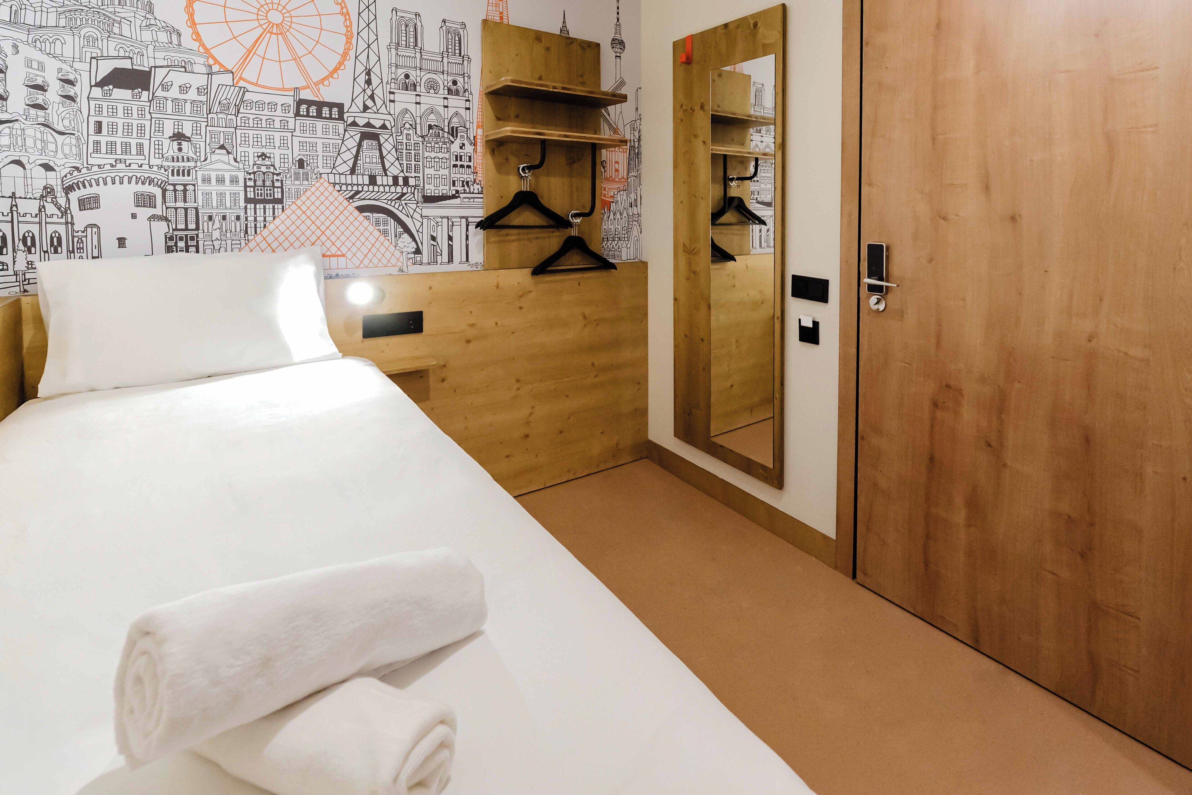 Una habitació d'hotel amb llit, una petita arca de fusta, un mirall i una paret amb un disseny de paisatge urbà.