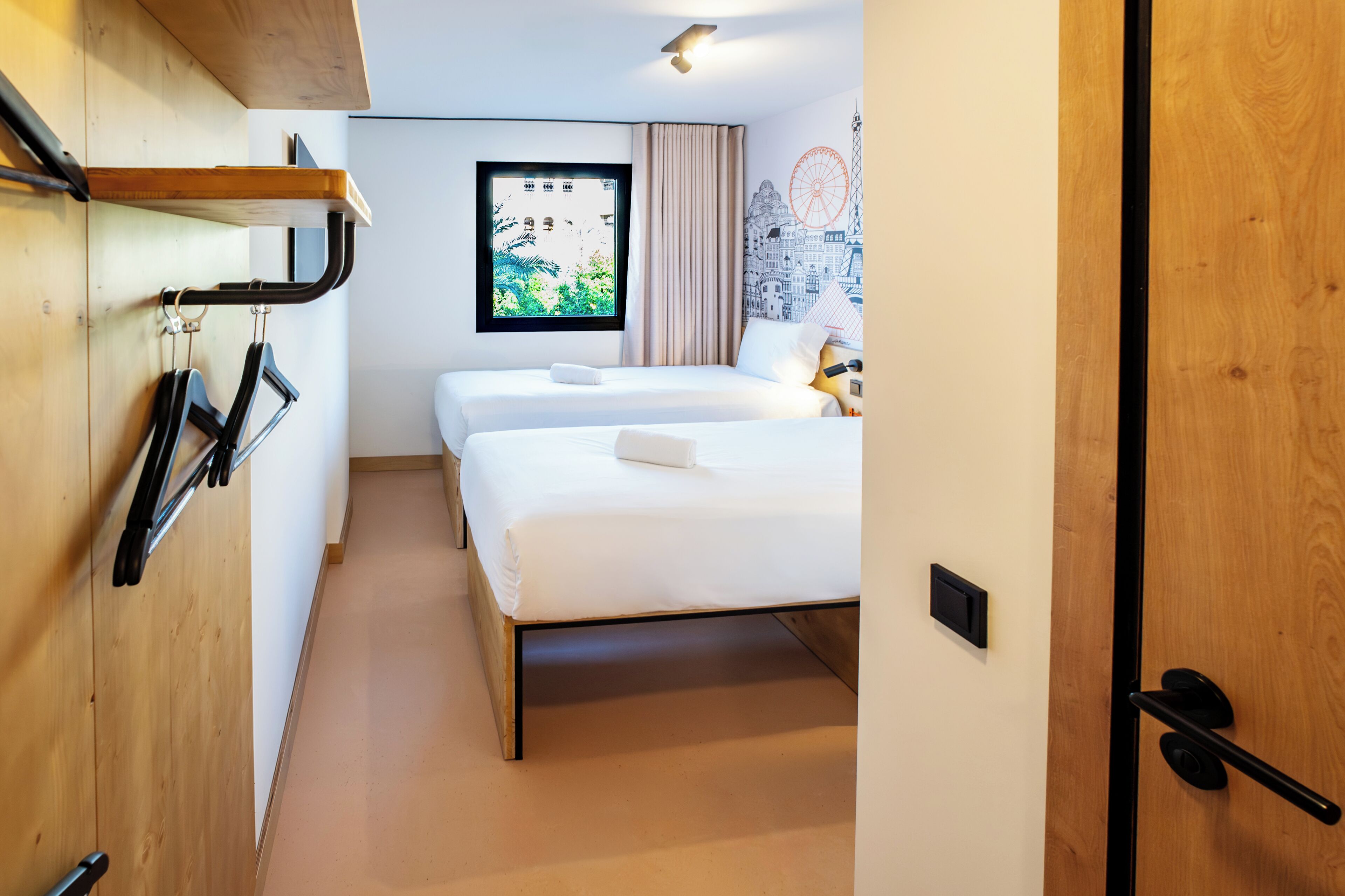 Ein Hotelzimmer mit Holzboden, zwei Betten, einem Fenster mit Blick auf die Stadt und einer Wandleuchte.
