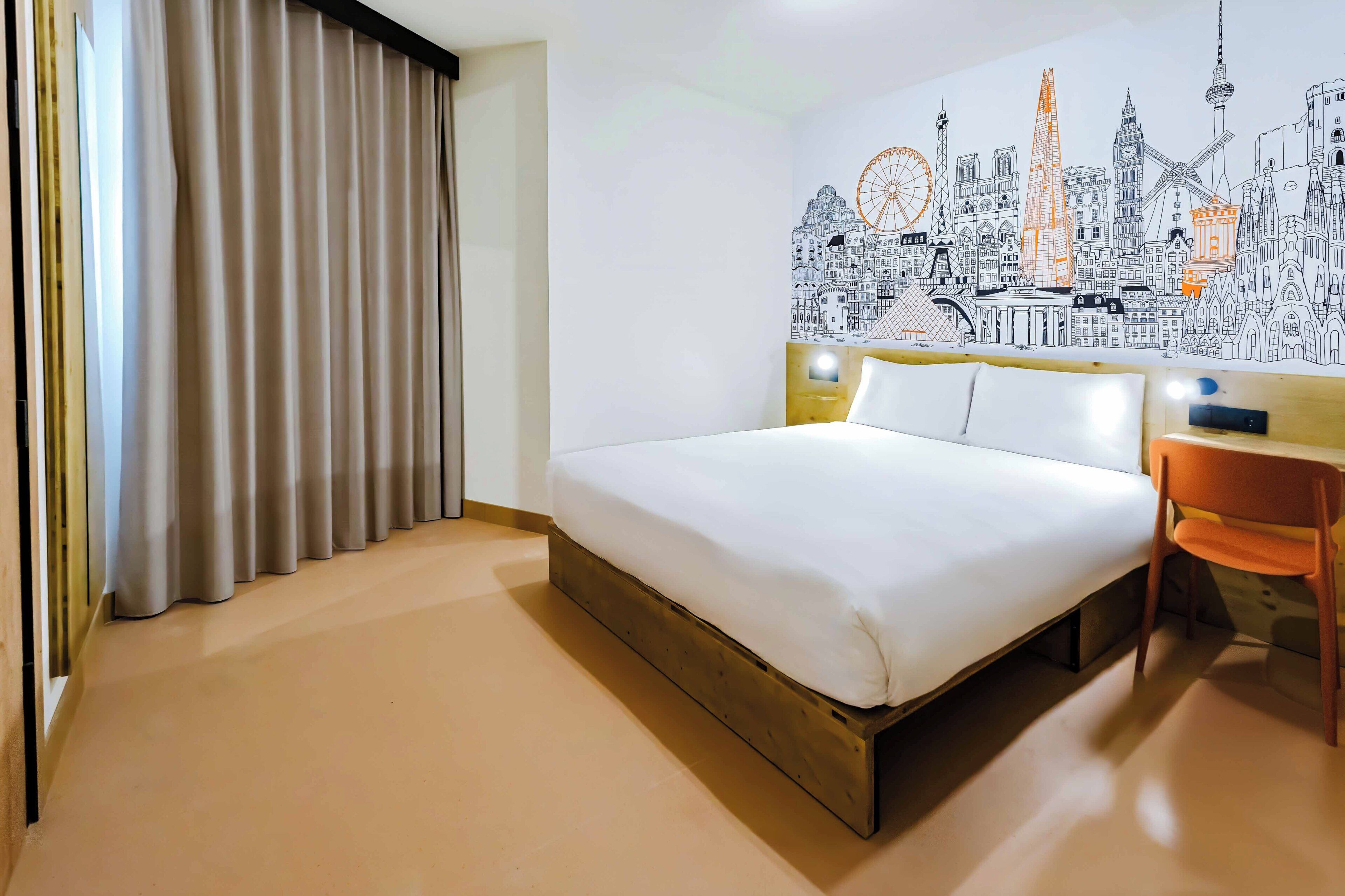 Habitación de hotel con cama, una silla, un mural en la pared y una ventana con cortinas.