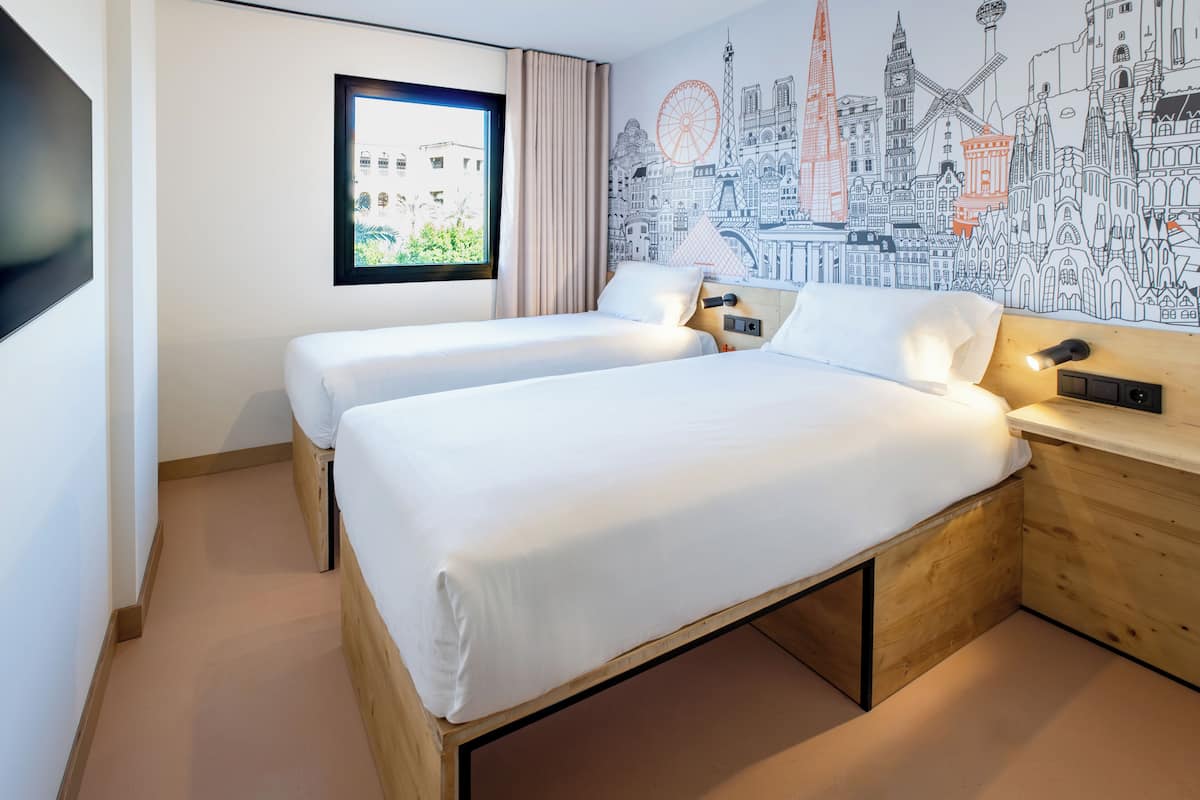 A hotel room with two beds, a large window, and a mural of a cityscape.