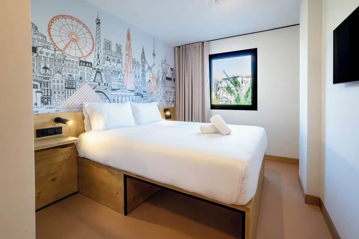 A modern hotel room with a large bed, a cityscape mural, a window with a view, and a flat-screen TV.