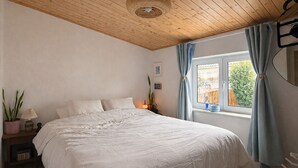 Un dormitorio con techo de madera, una cama grande, una mesita de noche con lámpara, una ventana con cortinas y vistas al exterior.