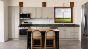 Uma cozinha moderna com armários brancos, uma geladeira de aço inoxidável, forno embutido e uma janela com vista para uma paisagem verdejante.