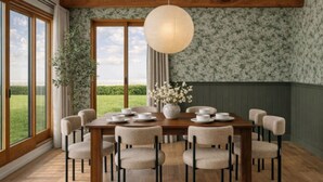Uma sala de jantar com uma mesa de madeira posta para oito pessoas, rodeada por cadeiras, com uma janela grande que oferece vista para o verde.