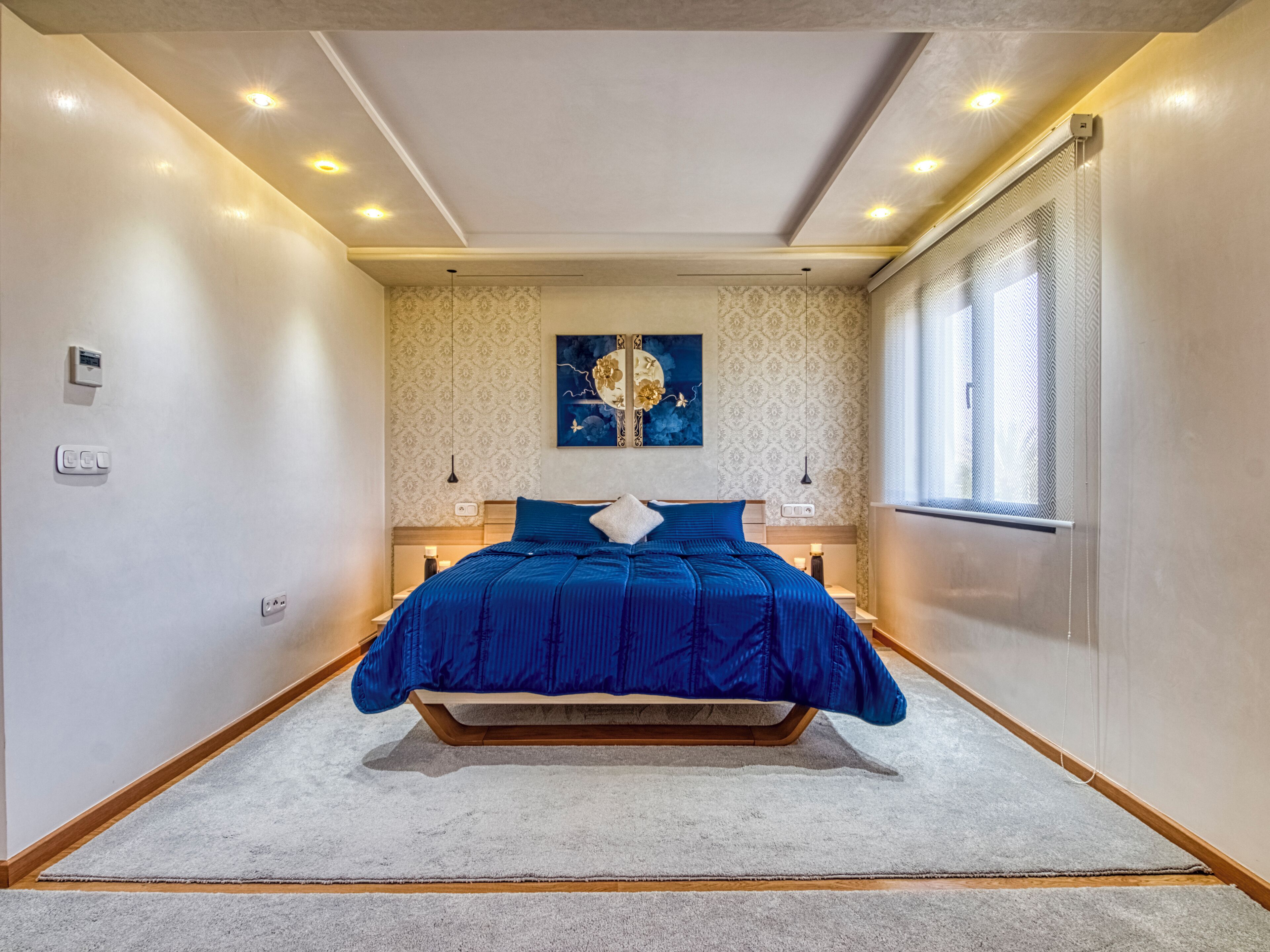 Une chambre avec un grand lit, un luminaire au plafond, un thermostat fixé au mur et une fenêtre avec des stores.
