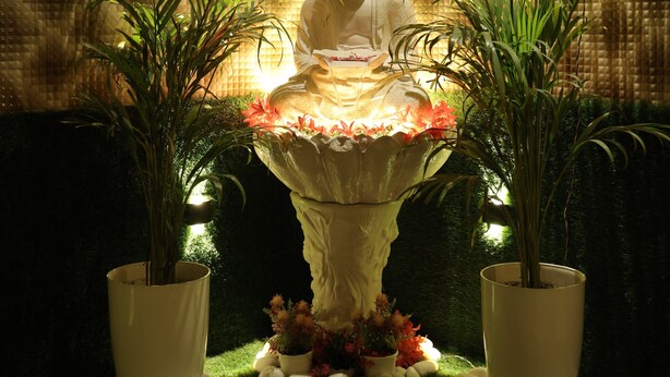 A Buddha statue in a meditative pose, surrounded by potted plants and a decorative fountain.