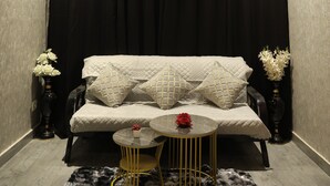 A room with a ceiling fan, black curtains, a sofa with cushions, a small round table with a vase of flowers, and a decorative rug.