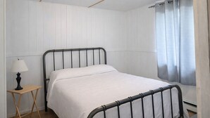 Una cama individual con estructura metálica, una mesita de noche con lámpara y una ventana con cortinas.