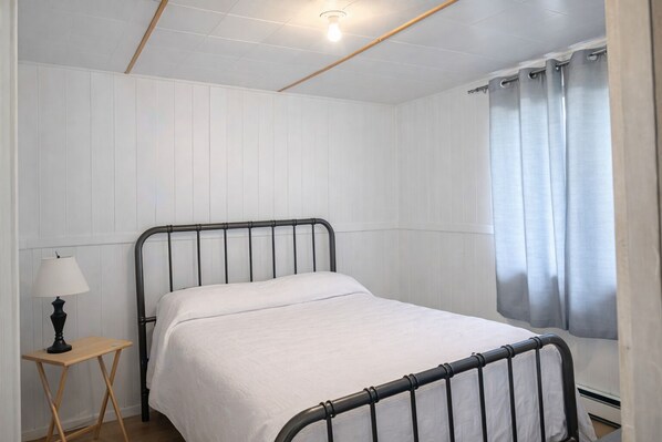 Una cama individual con estructura metálica, una mesita de noche con lámpara y una ventana con cortinas.