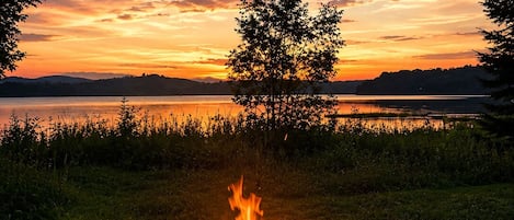 Nuotiolla palaa halkoja ja liekit, ja sen ympärillä on ruohoa ja kiviä. Taustalla näkyy puu ja järvi auringonlaskun värittämässä taivaassa.