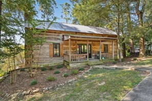 A rustic log cabin with a porch, surrounded by trees and a well-maintained lawn.