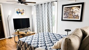 A bedroom with a bed, a TV, a ceiling fan, a window with curtains, and artwork on the walls.