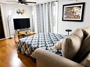 A bedroom with a bed, a TV, a ceiling fan, a window with curtains, and artwork on the walls.