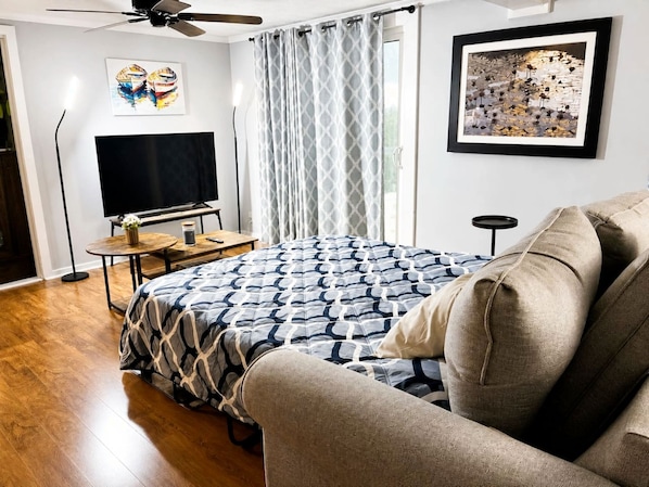 A bedroom with a bed, a TV, a ceiling fan, a window with curtains, and artwork on the walls.