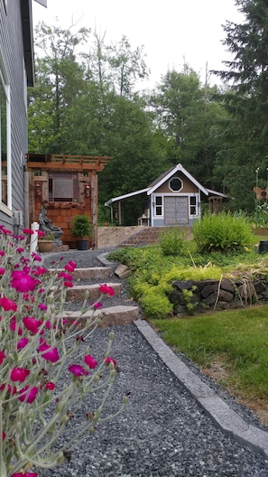 Ein Haus mit einer Holzveranda, einem kleinen Gartenhaus und einem Garten mit Kieswegen und rosa Blüten.
