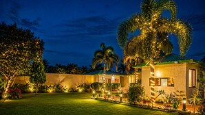 A well-lit resort with a manicured lawn, palm trees, and a building with a green roof. - Jungle Nest Retreat Resort (Ramnagar)