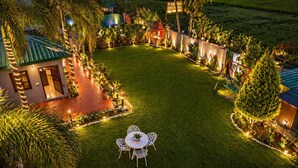 A well-lit outdoor area with a lawn, a small table with chairs, and a building with a green roof. - Jungle Nest Retreat Resort (Ramnagar)