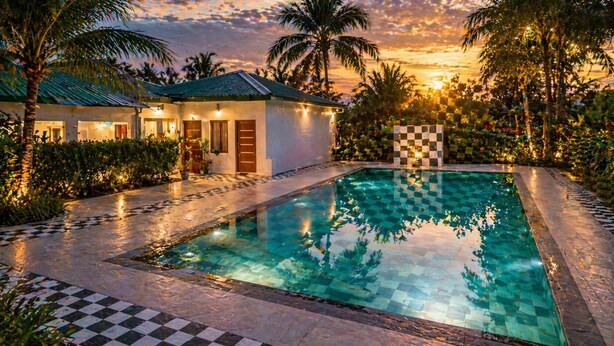 A swimming pool with a checkered border, surrounded by palm trees and a building with a tiled roof. - Jungle Nest Retreat Resort (Ramnagar)