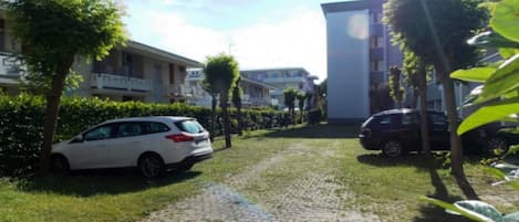 A paved pathway leading to a building with multiple balconies, surrounded by greenery and parked cars.