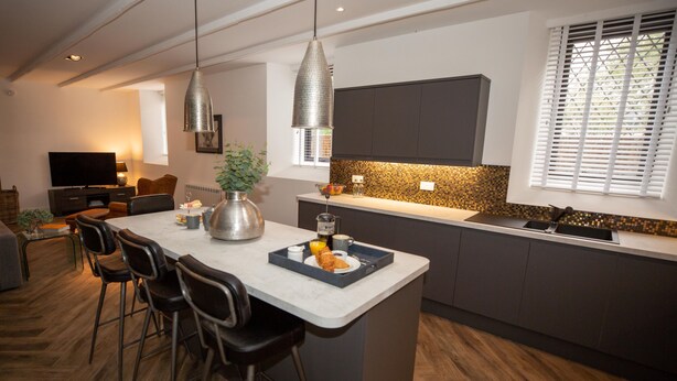 A modern kitchen with a central island, bar stools, a large TV, and a dining area with a sofa and chairs. - Stunning 3 Bed Converted Village Hall Near Kelso (Kelso)