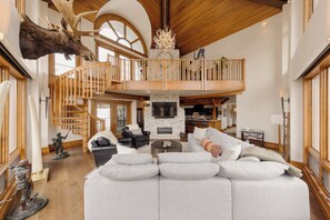 A spacious living area with a large white sectional sofa, a fireplace, wooden staircase, and a mounted moose head. - Premier Mountain Village Condo by InvitedHome | Hot Tub, Gym, Balcony Views (Telluride)