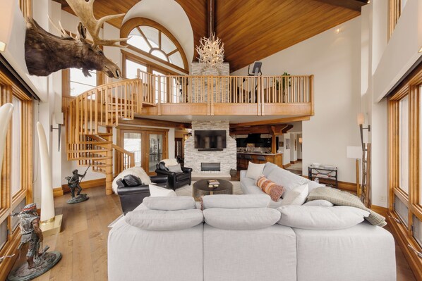 Een ruime woonkamer met een grote witte modulaire bank, een open haard, een houten trap en een opgehangen mooshoofd.