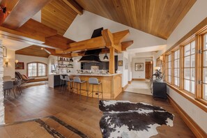 A spacious interior with a wooden ceiling, a bar area with stools, a dining table, and a large window. - Premier Mountain Village Condo by InvitedHome | Hot Tub, Gym, Balcony Views (Telluride)