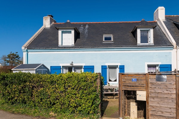 Una casa de dos pisos con fachada azul, una ventana pequeña y una puerta con un persiano azul.