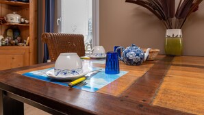 Ein Esstisch mit Platzdeckchen, einer blauen Glasgießkanne, einer weißen Teekanne und einem Korbstuhl.