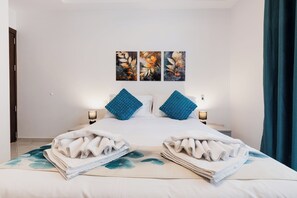 Una cama bien tendida con toallas dobladas, dos almohadas azules y tres cuadros colgados en la pared.