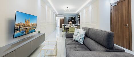 A modern living room with a grey sofa, a flat-screen TV mounted on the wall, and a dining area in the background.
