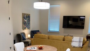 A modern living room with a round wooden table, white chairs, a yellow sofa, a ceiling fan, and a mounted TV. - Beautiful New Construction 2 - Bedroom / 2 Bath located in West University. (Austin)